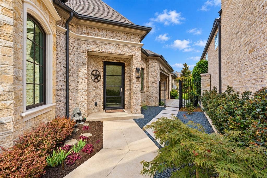 6816 Cousteau Court, Plano, TX 75024 Elegant brick courtyard entry with professional hardscape paving, mature landscaping including ornamental shrubs and evergreens, arched architectural detailing, and dark-framed entry door with sidelights