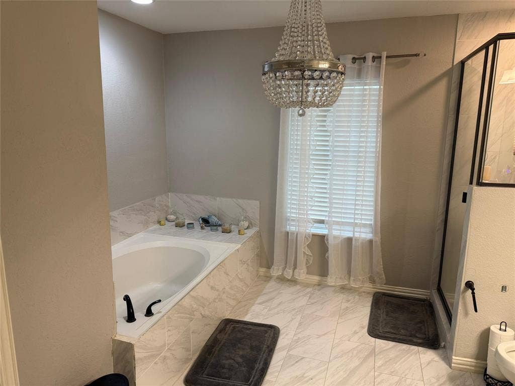 Built-in soaking tub with black fixtures, tiled surround, white window blinds, and crystal chandelier