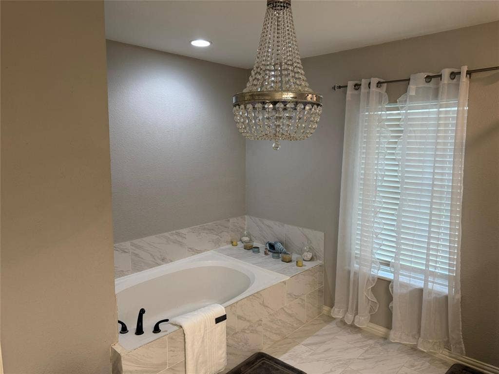 Recessed soaking tub with surrounding marble-look tile, crystal chandelier, and matching marble-look tile flooring