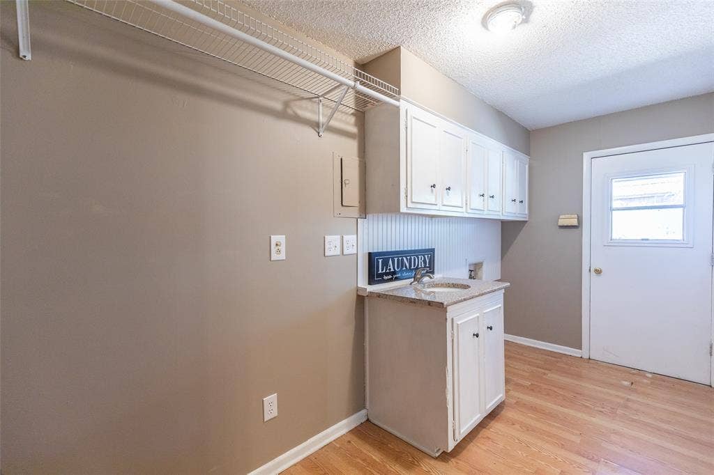Laundry area featuring wood-finish flooring, a utility sink with a stone-look countertop, white cabinetry, overhead wire shelving, and a white exterior door