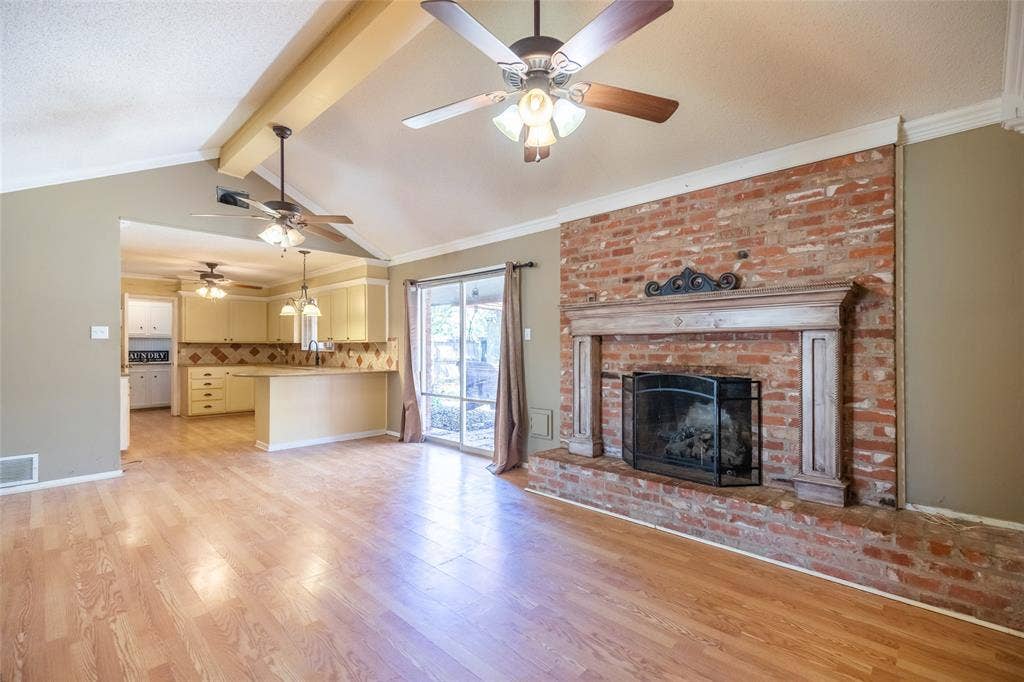 Vaulted ceiling with exposed beams, wood-finish flooring, brick fireplace with wood mantel, sliding glass door, and recessed kitchen with built-in cabinetry