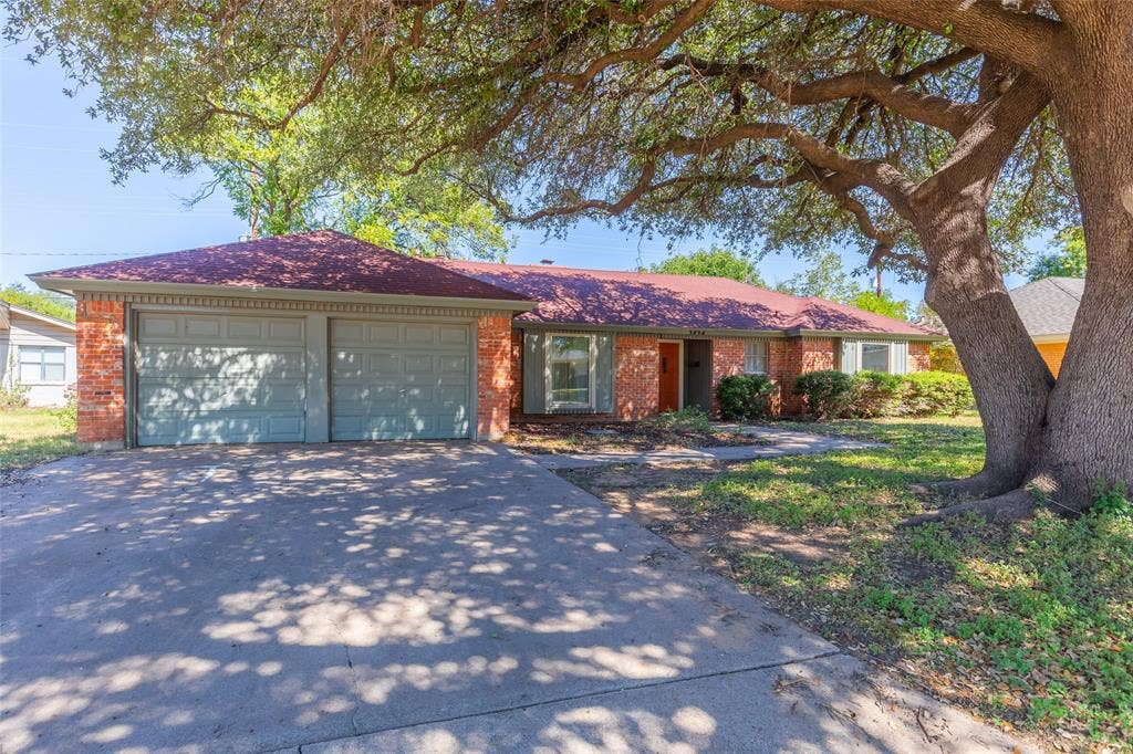 Brick exterior with a two-car garage, red shingle roof, and mature trees providing natural shade