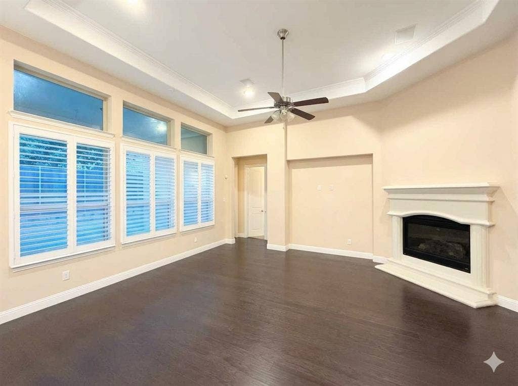 5917 Trantina Lane, Frisco, TX 75035 Tray ceiling with crown molding, dark wood-finish flooring, white plantation shutters, electric fireplace with white mantel, and ceiling fan