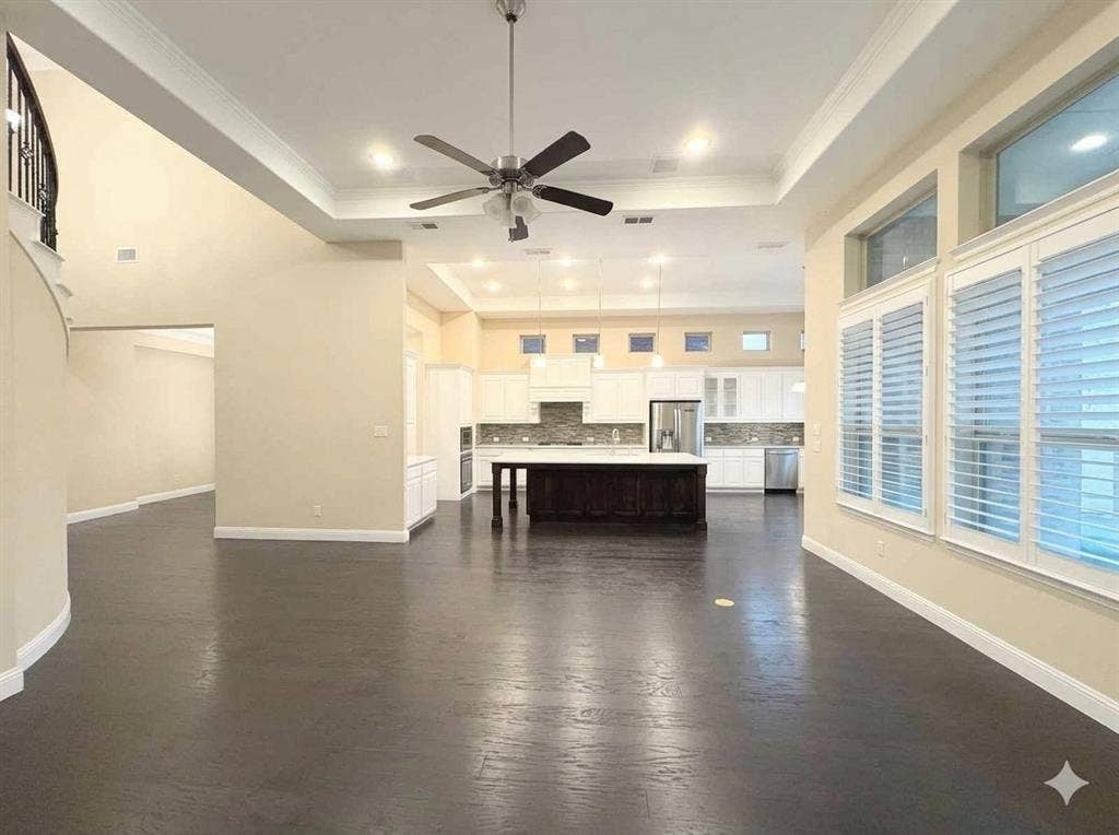 5917 Trantina Lane, Frisco, TX 75035 Spacious living area featuring dark wood-finish flooring, a tray ceiling with a ceiling fan, and large windows with plantation shutters