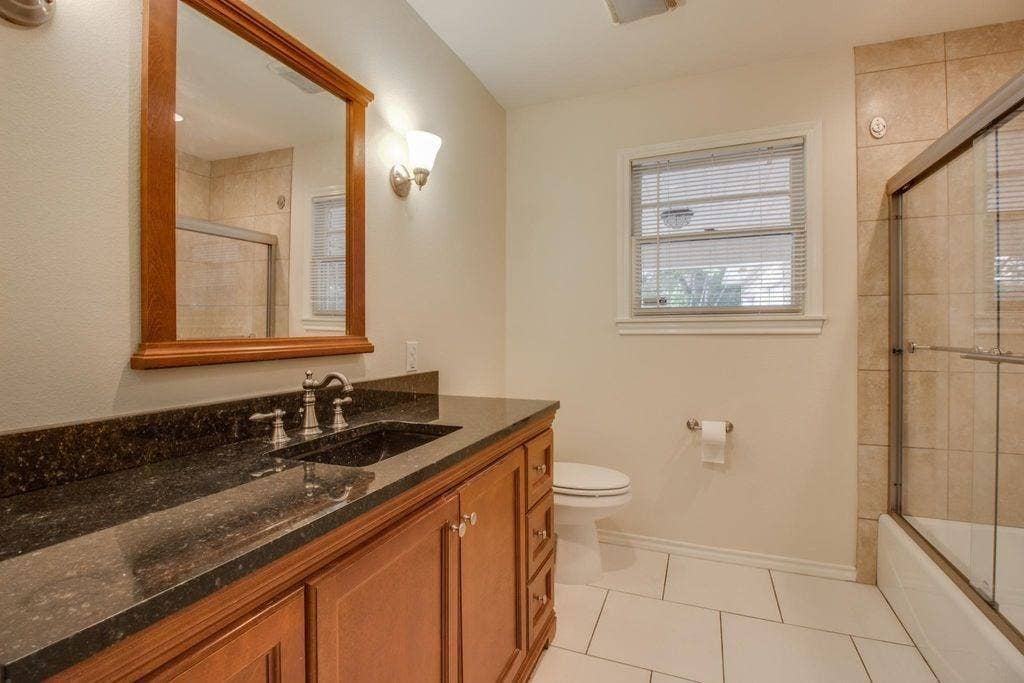 Dark-toned vanity with dual sinks and a light-toned countertop, framed mirror, wall-mounted light fixtures, tile flooring, and a tiled shower with a glass enclosure