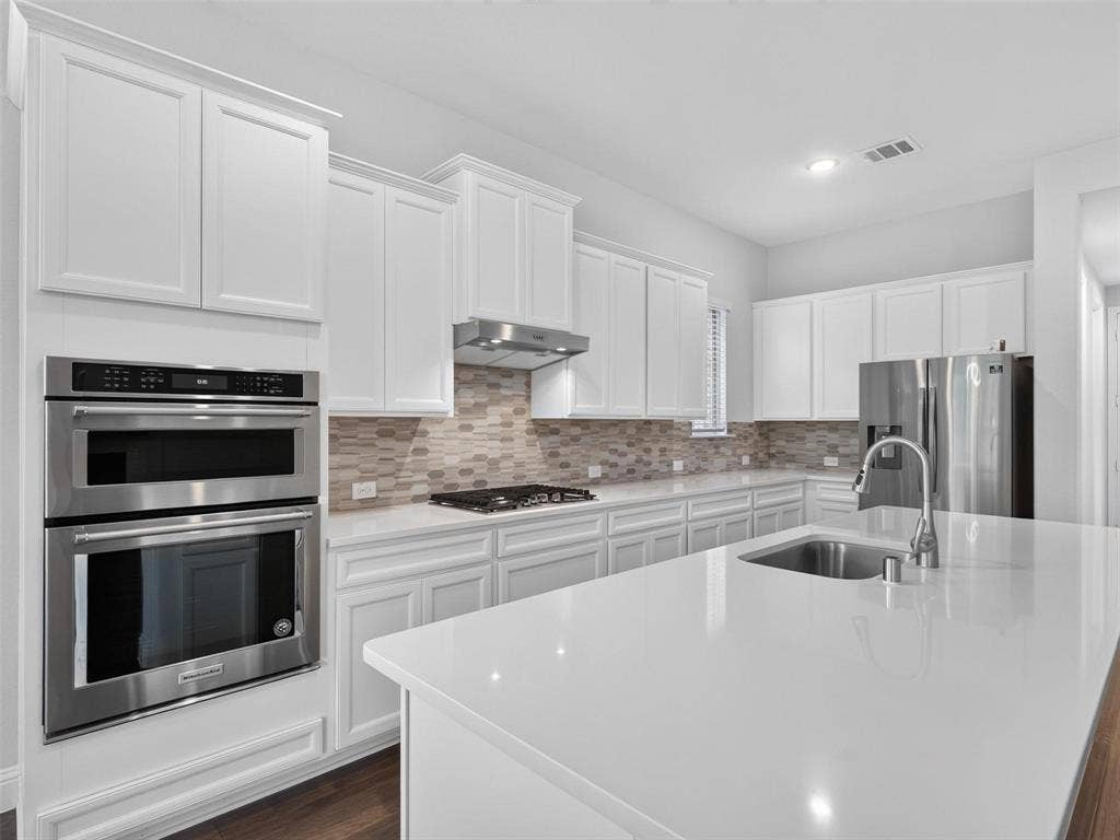 6504 Dynamite Drive, McKinney, TX 75070 White shaker-style cabinetry, stainless steel appliances, light-toned tiled backsplash, white countertop surfaces, and a central island with an undermount sink