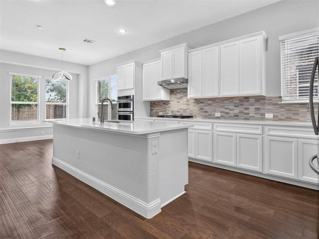 6504 Dynamite Drive, McKinney, TX 75070 Expansive kitchen island with a quartz countertop and integrated sink, stainless steel appliances, white cabinetry, decorative tile backsplash, and wood-finish flooring