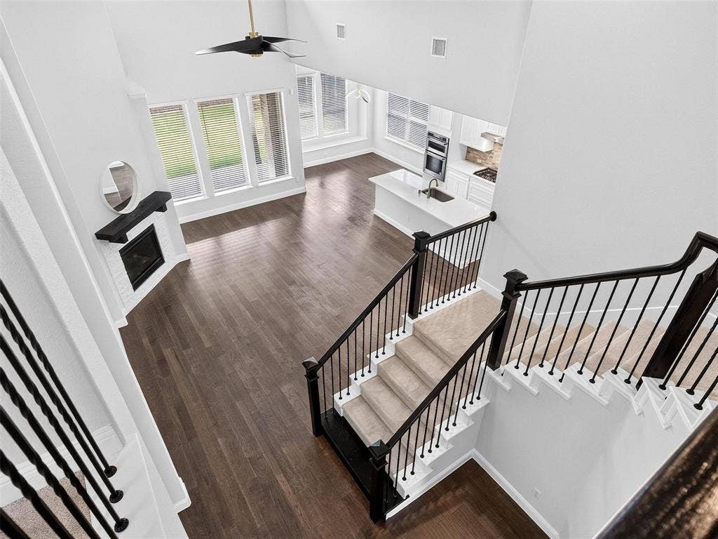 6504 Dynamite Drive, McKinney, TX 75070 Vaulted ceiling with a ceiling fan, wood-finish flooring, white brick fireplace with a dark mantel, white kitchen island with a sink, and a carpeted staircase with dark railings