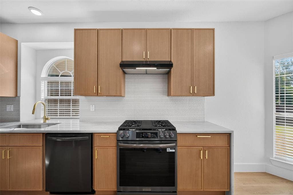 705 N Jordan Road, McKinney, TX 75071 Light wood-finish cabinetry with gold-tone hardware, white countertops, black gas range with matching range hood, and a built-in black dishwasher