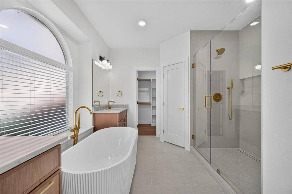 705 N Jordan Road, McKinney, TX 75071 Freestanding soaking tub with floor-mounted gold-finish faucet, walk-in shower with glass enclosure, dual vanity with undermount sinks, and arched window with privacy blinds
