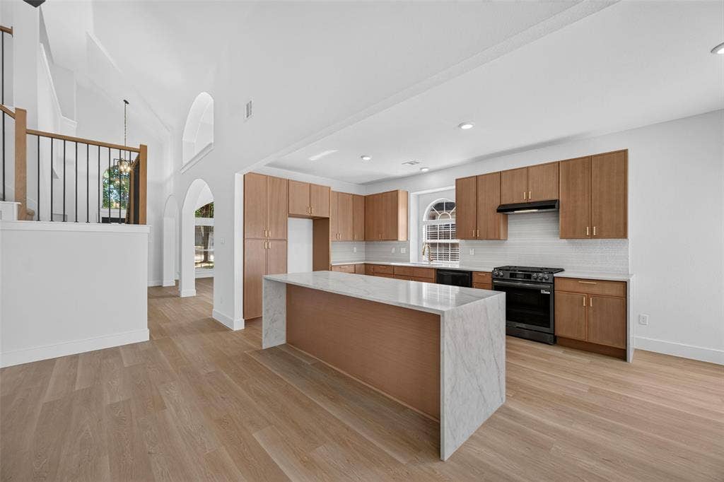 705 N Jordan Road, McKinney, TX 75071 Spacious kitchen featuring an expansive island with a waterfall countertop, wood-finish cabinetry, black appliances, and a tiled backsplash