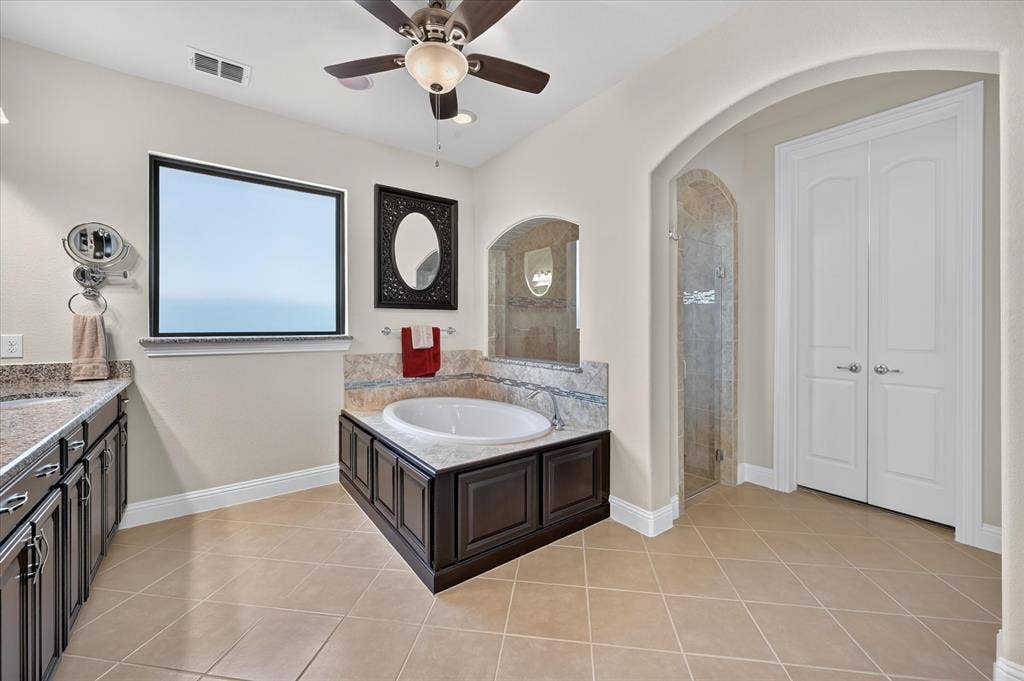 Built-in soaking tub, dual vanity with stone countertops, arched doorway, tiled shower, and tiled flooring