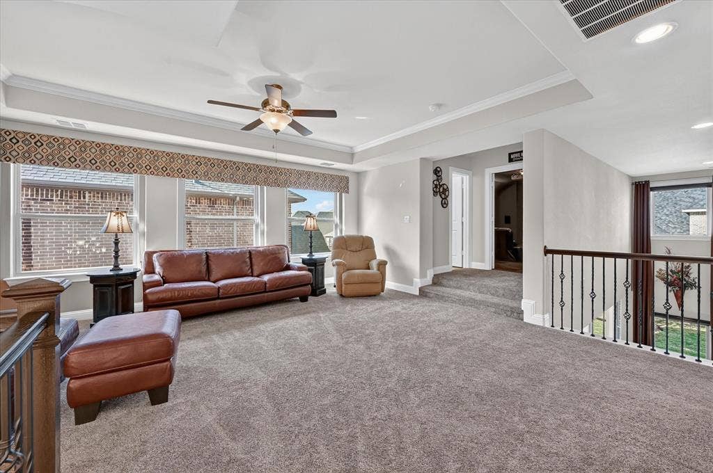Spacious carpeted room featuring a ceiling fan, tray ceiling, multiple windows, and a black wrought iron railing