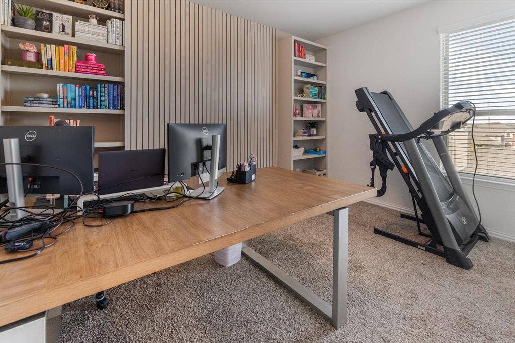 3605 Barby Court, Midlothian, TX 76065 Modern home office featuring a spacious wood-finish desk, built-in shelving with vertical slat wall paneling, dual monitor setup with ergonomic workspace, natural light from large windows with blinds, and neutral carpeting throughout