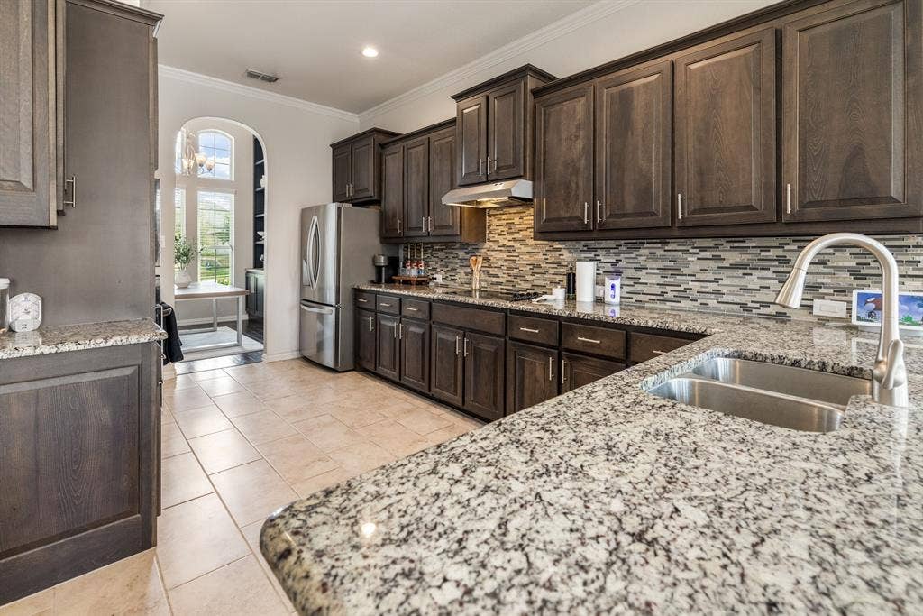 3605 Barby Court, Midlothian, TX 76065 Kitchen featuring extensive dark wood cabinetry, granite countertops, stainless steel appliances, a double basin sink with a high-arch faucet, and a mosaic tile backsplash