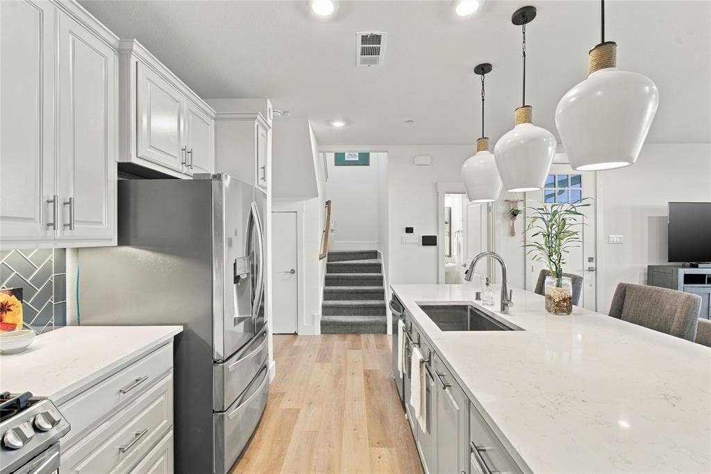 Kitchen featuring white cabinetry, an expansive island with a light-toned countertop, stainless steel appliances, herringbone-patterned tile backsplash, and three modern pendant light fixtures