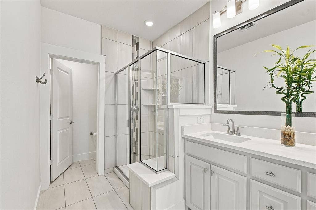 Bathroom featuring white cabinetry, solid-surface countertop, walk-in shower with vertical decorative tile inlay, and a large-format framed mirror with three-light fixture