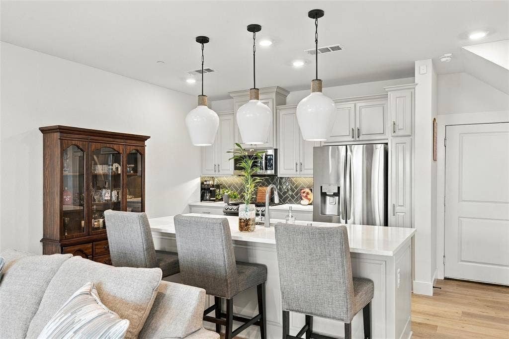 Kitchen featuring white shaker-style cabinetry, a quartz-topped island with a stainless steel undermount sink, a dark tile herringbone backsplash, and three oversized glass pendant lights