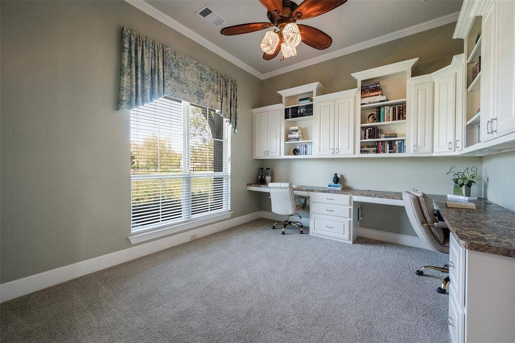 Built-in white cabinetry with extensive shelving, stone-surface countertop, multi-drawer desk workspace, ceiling fan with light fixture, and large window with blinds