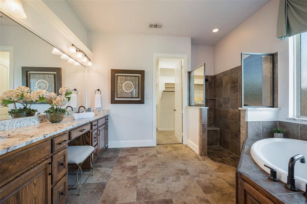 Dual-sink vanity with stone-finish countertop, walk-in shower with tile surround, deep soaking tub, and tile flooring
