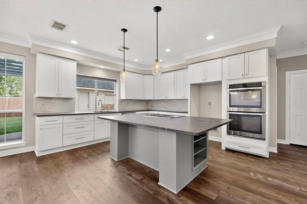 Spacious kitchen featuring white cabinetry, wood-finish flooring, a central island with integrated shelving, stainless steel double ovens, and recessed lighting