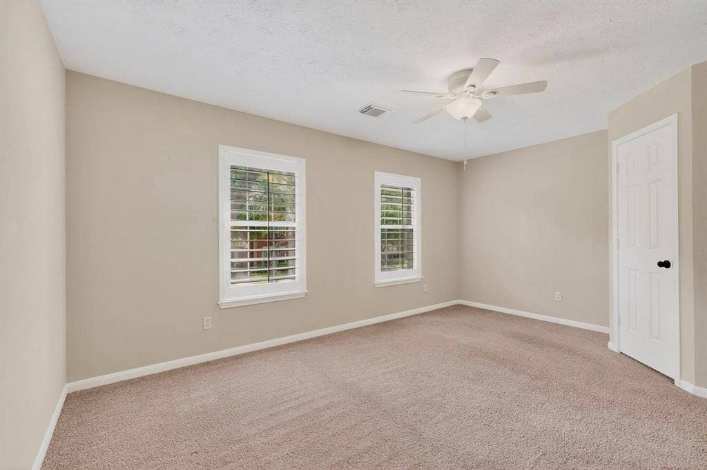 Neutral wall tones, carpeted flooring, two windows with white plantation shutters, white ceiling fan with integrated lighting, and a white paneled door with black hardware