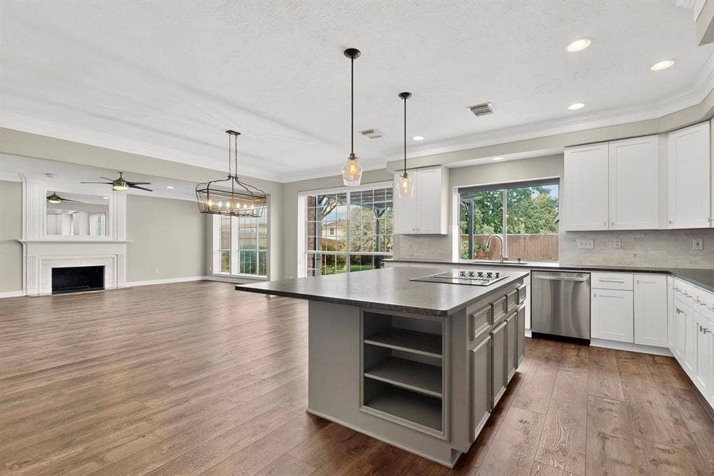 Open-concept living space featuring wood-finish flooring, a kitchen island with built-in cooktop, white shaker-style cabinetry, and a fireplace with a mantel
