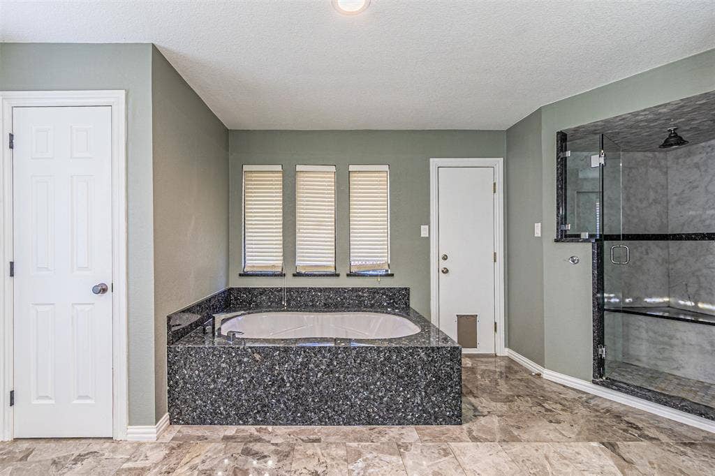8808 Sylvan Court, Fort Worth, TX 76120 Soaking tub with a dark stone surround, three windows with blinds, glass-enclosed shower with a dark stone base, and tiled flooring