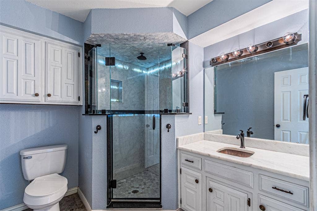 8808 Sylvan Court, Fort Worth, TX 76120 Glass-enclosed shower with a rainfall showerhead, vanity with an integrated sink, broad wall mirror, and white shaker-style cabinetry