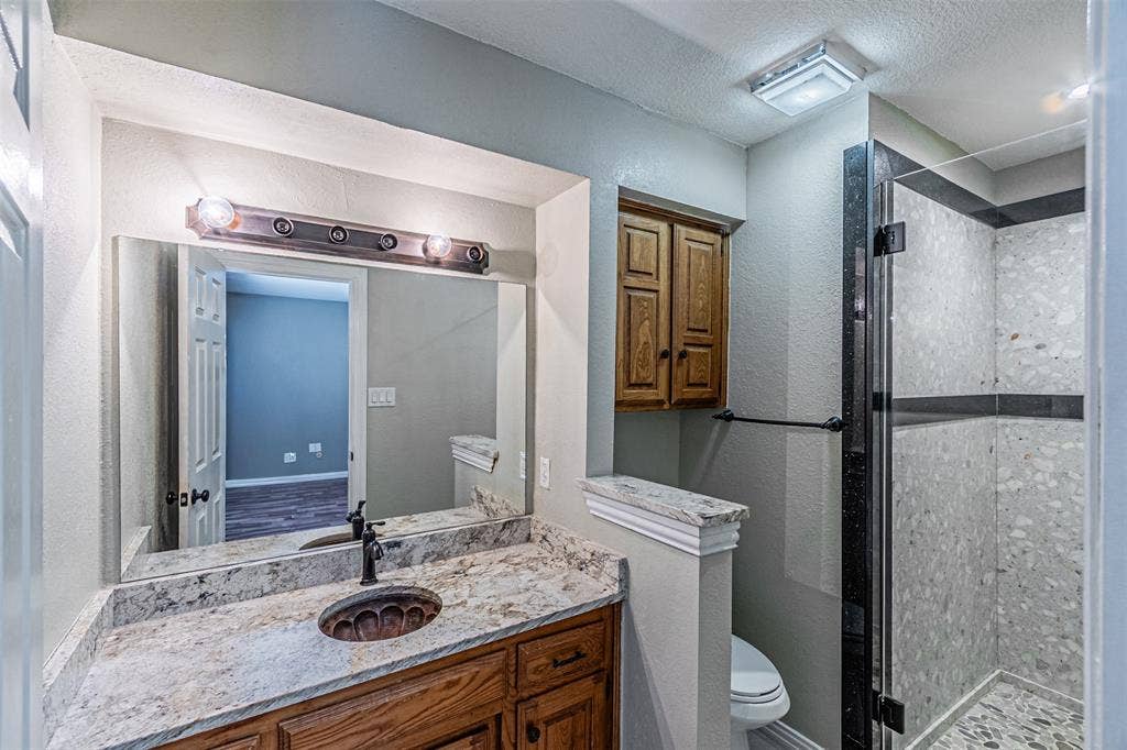 8808 Sylvan Court, Fort Worth, TX 76120 Vanity with an integrated sink, granite-style countertop, wood-finish cabinetry, and a glass-enclosed shower featuring tiled walls