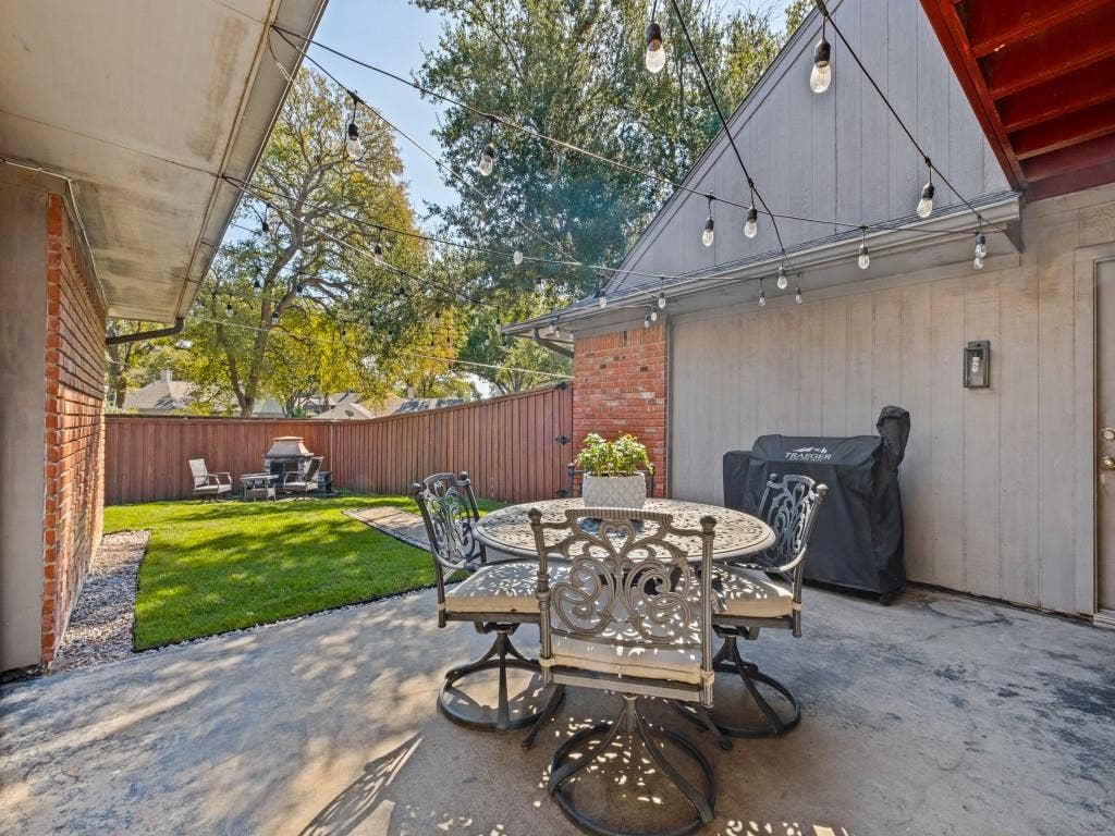 5730 Winding Woods Trail, Dallas, TX 75227 Backyard patio featuring red brick accents, vertical siding, an attached wall-mounted lantern, and a wood privacy fence enclosing a lawn area with mature trees
