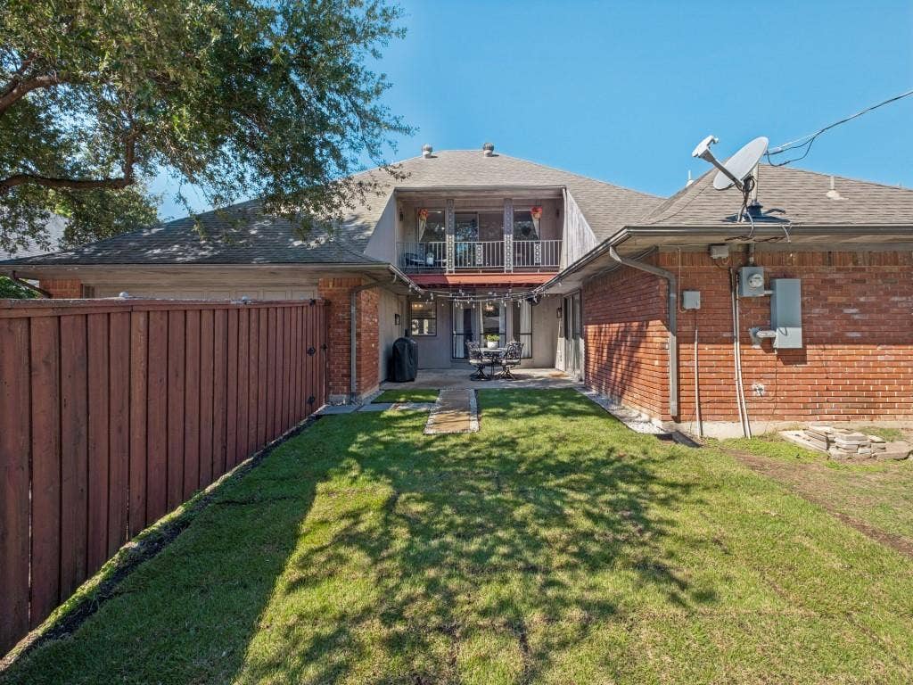 5730 Winding Woods Trail, Dallas, TX 75227 Brick exterior with a stone-paved patio, wooden privacy fencing, upper-level balcony with metal railing, and a grass lawn