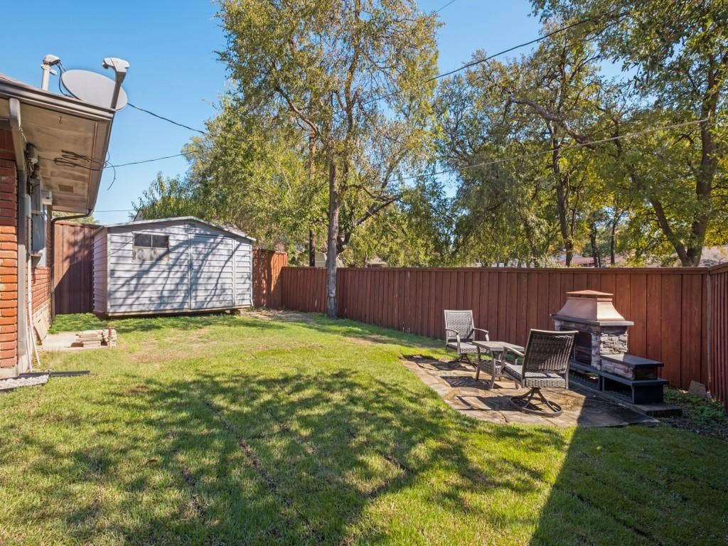5730 Winding Woods Trail, Dallas, TX 75227 Backyard featuring a grass lawn, stone patio, stone-accented outdoor fireplace, storage shed, and wood-panel fencing