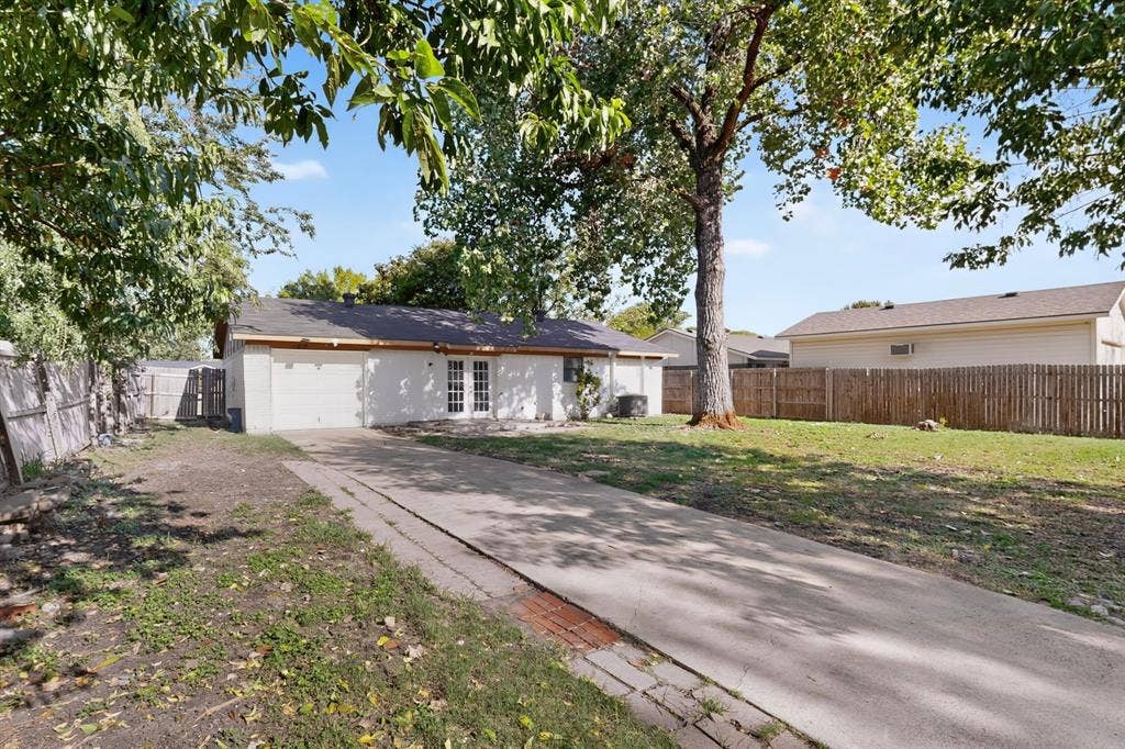 1512 Bay Shore Drive, Garland, TX 75040 Exterior rear facade featuring a concrete driveway, multi-pane glass patio doors, mature trees, and a wood privacy fence