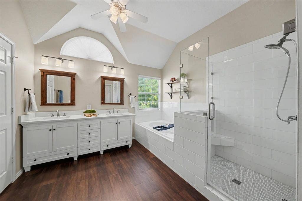 Vaulted ceiling with an arched window, dual vanity with white cabinetry and integrated sinks, walk-in shower with glass enclosure, white subway tile surround, and mosaic tile flooring, and a built-in soaking tub