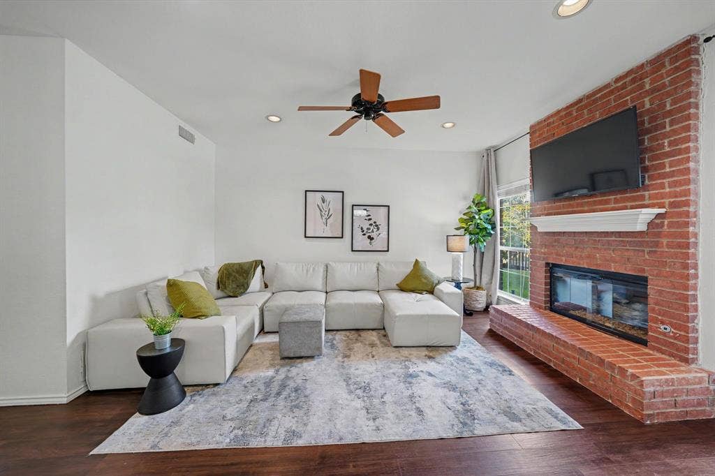 Living area featuring wood-finish flooring, a red brick fireplace with a white mantel, and a ceiling fan with integrated lighting