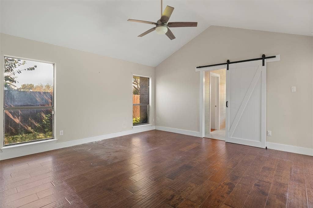 Vaulted ceiling with a ceiling fan, wood-finish flooring, two large windows, and a white barn door
