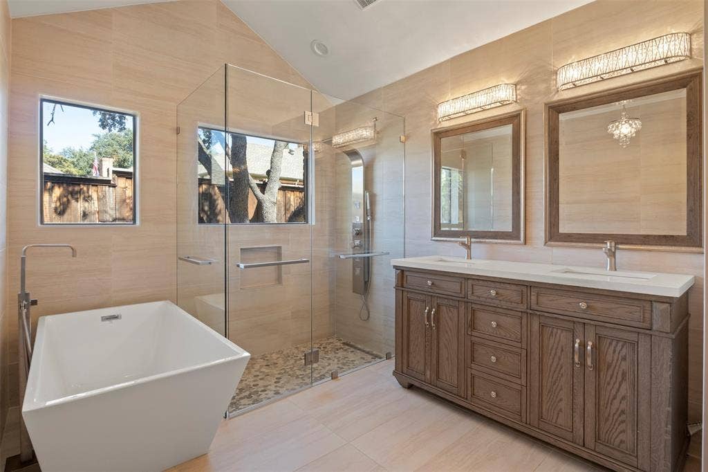 Freestanding soaking tub with floor-mounted faucet, glass-enclosed shower with pebble floor, dual vanity with wood-finish cabinetry, and framed mirrors with sconce lighting