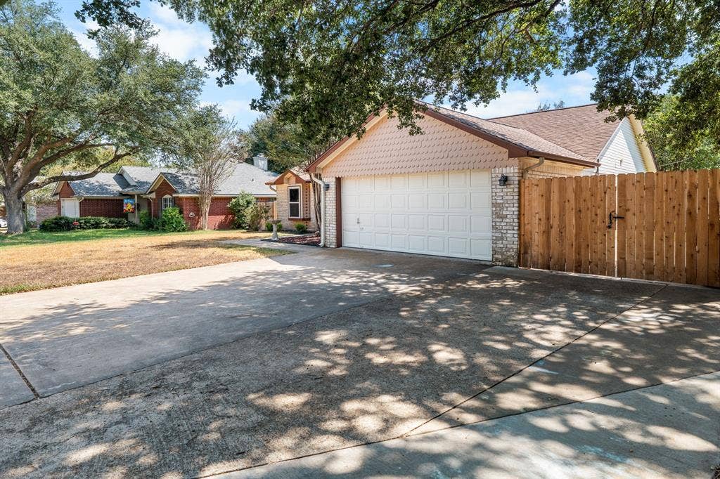 10712 Apache Drive, Waco, TX 76712 Expansive concrete driveway leading to an attached garage with a panel door, complemented by a wooden fence with a gate, and a brick veneer exterior