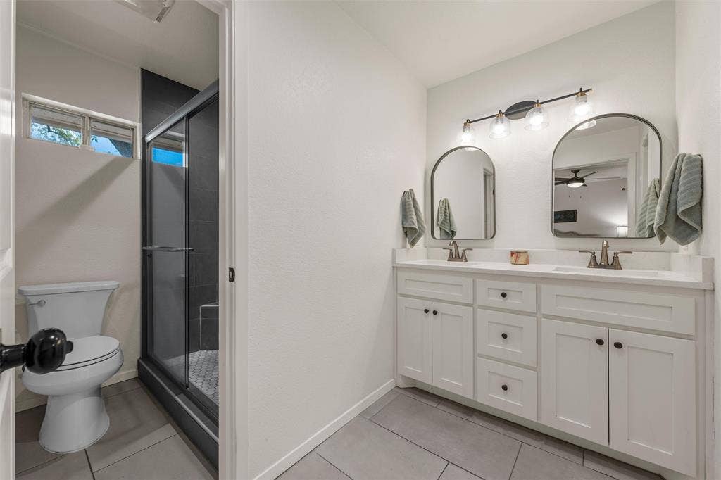 1712 Laurel Lake Drive, Waco, TX 76710 Dual vanity with white cabinetry and integrated sinks, a walk-in shower with dark-tiled walls, a toilet, and gray tile flooring