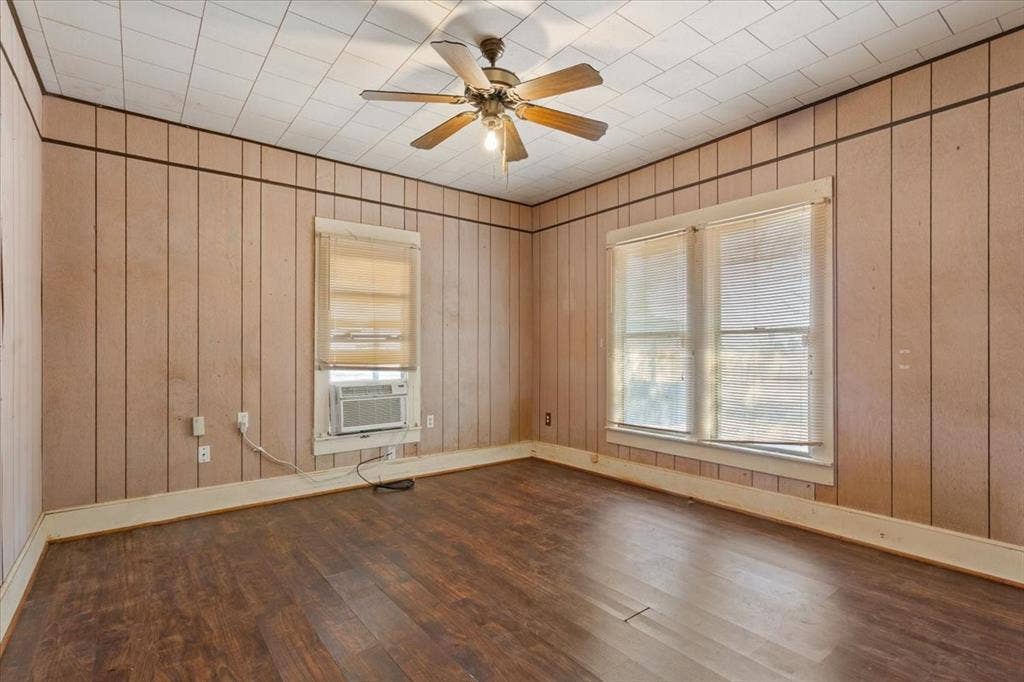 4246 Old Caufield Ranch Road, McGregor, TX 76657 Spacious corner room with wood-finish flooring, vertical wood paneling throughout, ceiling fan with brushed metal finish, and dual windows with roller shades providing natural light