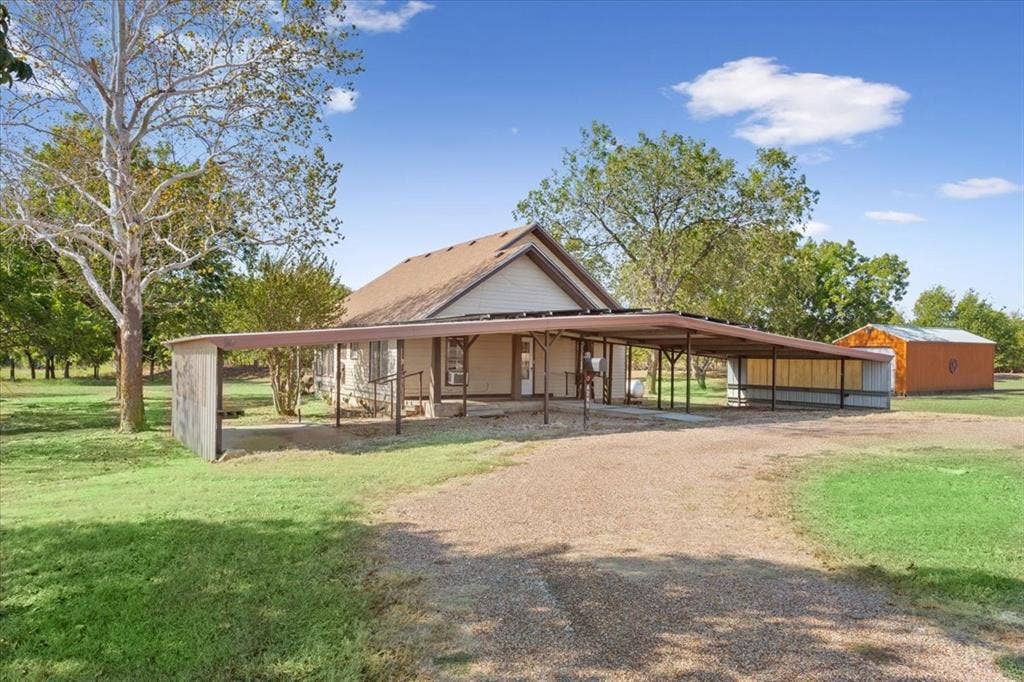 4246 Old Caufield Ranch Road, McGregor, TX 76657 Gable roof structure with a covered porch entry, expansive gravel driveway, long attached carport, and a detached outbuilding