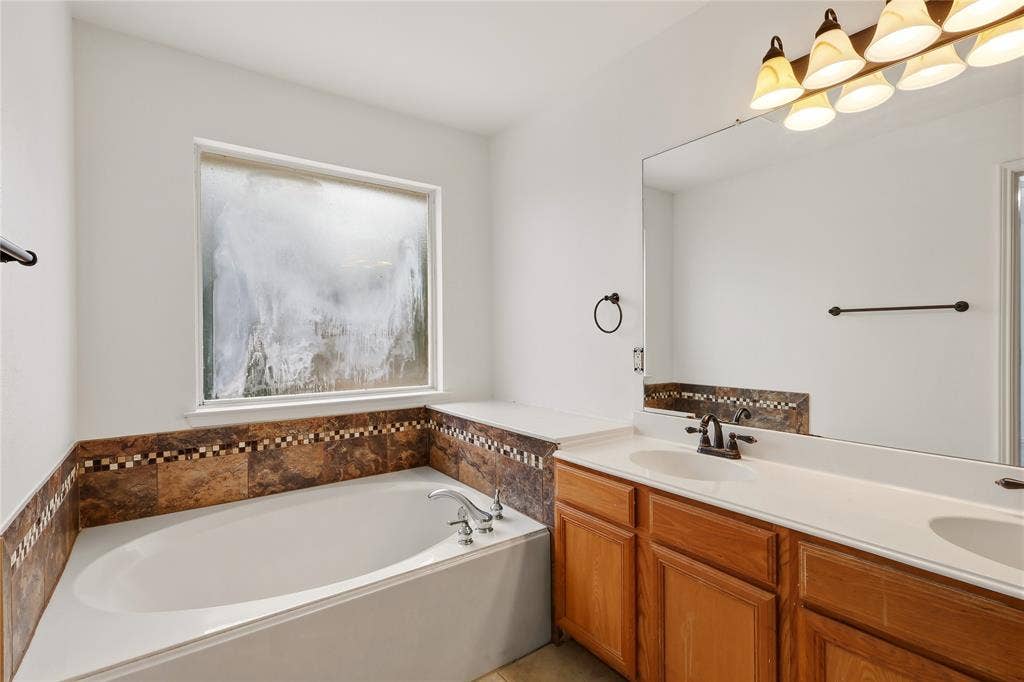 9105 Norman Drive, Plano, TX 75025 Built-in soaking tub, dual vanity with wood-finish cabinetry, extensive vanity mirror, wall-mounted light fixture with five sconces, and a tiled backsplash and tub surround