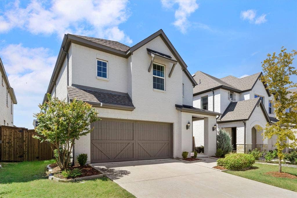 1456 Kit King Drive, Little Elm, TX 75036 White brick exterior with dark trim accents, concrete driveway, two-car garage with wood-finish doors, and mature landscaping