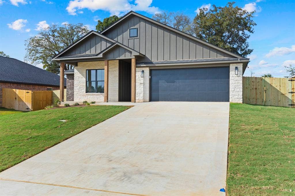 16514 Crossing Circle, Lindale, TX 75771 Contemporary exterior featuring vertical siding and stone accents, a deep gray garage door, a concrete driveway, wood support beams, and a wood privacy fence