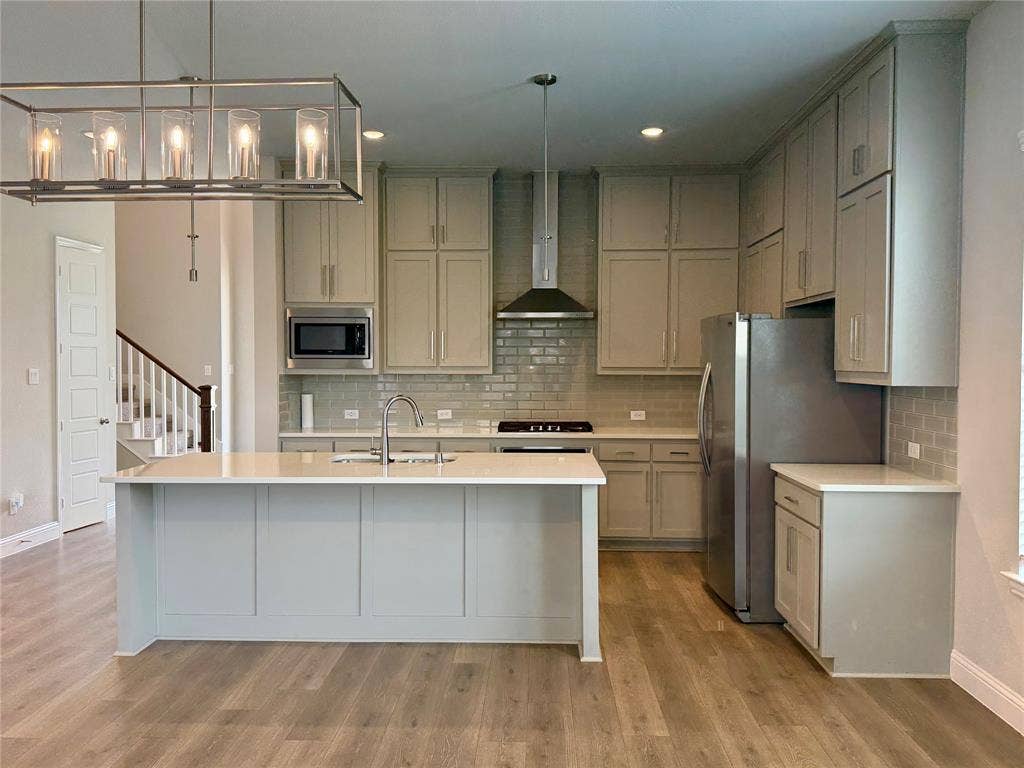 1196 Putnam Drive, Frisco, TX 75036 Kitchen featuring a large center island with a sink, light-toned cabinetry, stainless steel appliances, subway tile backsplash, and wood-finish flooring
