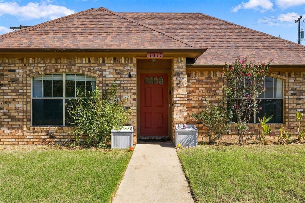 1015 Shelly Drive, Keene, TX 76031 Brick exterior with arched window detailing, a red entry door with decorative glass, and a brown shingle roof