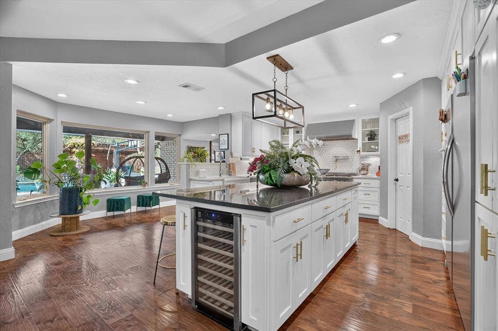 7016 Battle Creek Road, Fort Worth, TX 76116 Spacious kitchen featuring an expansive center island with a built-in beverage refrigerator, wood-finish flooring, recessed lighting, white cabinetry with gold-tone hardware, and a tile backsplash