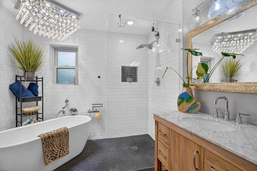 7016 Battle Creek Road, Fort Worth, TX 76116 Freestanding soaking tub, walk-in shower with clear glass enclosure, white subway tile walls, wood-finish vanity with stone countertop, and crystal chandelier fixture