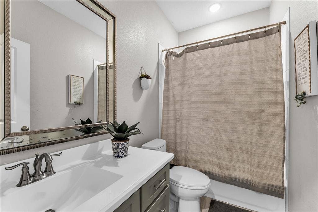 Spa-inspired bathroom featuring a framed mirror with integrated lighting, white ceramic sink with polished chrome faucet and handles, white subway tile surround, neutral linen shower curtain with metal rod, white porcelain toilet, and wood-finish vanity cabinetry with ample storage